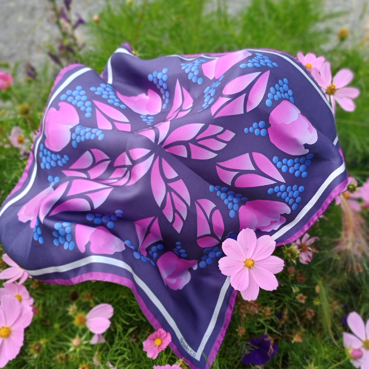 A square silk scarf with a pink and blue floral pattern on a dark purple background, styled outdoors on green grass among pink flowers.