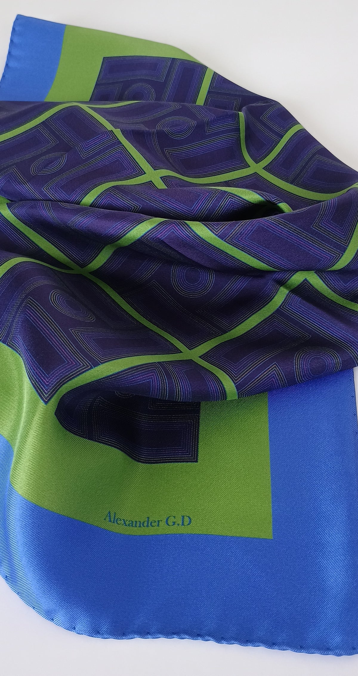 Blue and green patterned fabric with 'Alexander G.D.' branding on a white background with day light showing the sheen of the silk