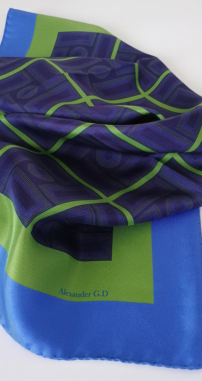 Blue and green patterned fabric with 'Alexander G.D.' branding on a white background with day light showing the sheen of the silk