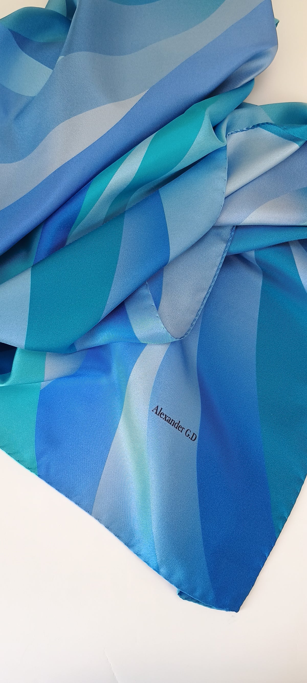 A folded blue scarf with a  sheen of silk, featuring shades of royal blue, cyan, and light blue, with the brand name Alexandra G.D printed in black near the corner.